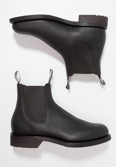 Black chelsea boots featuring smooth leather, elastic side panels, pull tabs, and a low, stacked wooden heel. Flat sole with a textured finish.