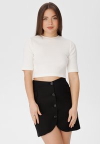 Bílý žebrovaný crop top s krátkým rukávem doplněný černou sukní vysokého pasu s vykrojeným lemem a šesti černými knoflíky.