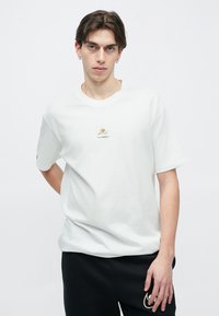 Fehér pamut póló laza fazonban, a mellkason kis arany hímzéssel díszítve. Rövid ujjú, kerek nyakkivágással.