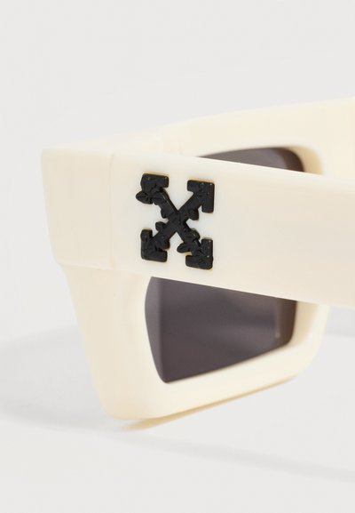 Gros plan du bras beige de lunettes de soleil avec logo flèche noire et lentille rectangulaire foncée visible en arrière-plan.