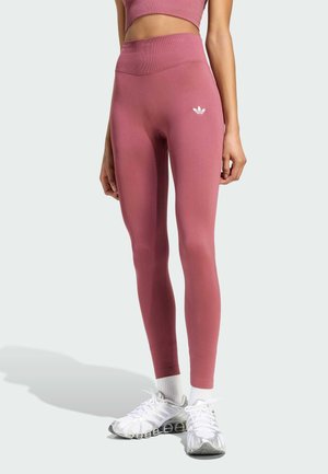 Frau trägt hochgeschnittene, mauvefarbene Leggings und passendes Crop-Top mit weißem Adidas-Logo, weiße Socken und weiße Sportschuhe.