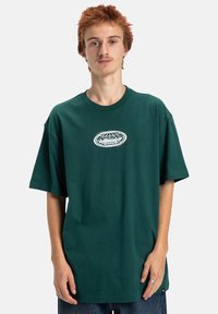 Zielona bawełniana koszulka z krótkimi rękawami, okrągłym dekoltem i centralnym białym logo graficznym. Luźny krój z miękką teksturą.