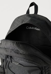 Μαύρο σακίδιο Calvin Klein με φερμουάρ μερικώς ανοιχτό, που δείχνει το εσωτερικό λογότυπο και το υφασμάτινο μοτίβο εξωτερικά.