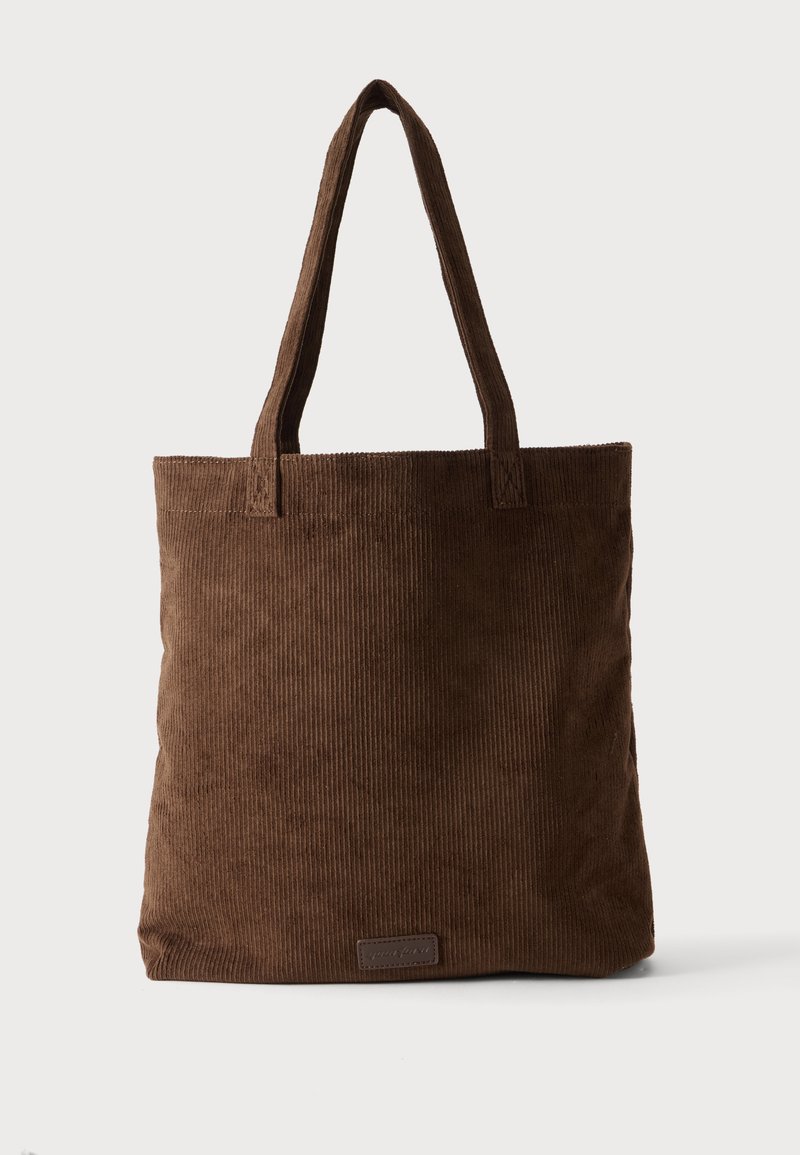 Sac cabas en velours côtelé marron avec deux longues poignées et un petit patch rectangulaire près du bas au centre.