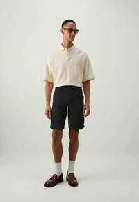 Gai bēšs īsām piedurknēm polo krekls ar apkakli, apvienots ar tumšiem šortiem, potīšu zeķēm un brūnām kurpēm. Aksesuāri ietver saulesbrilles.