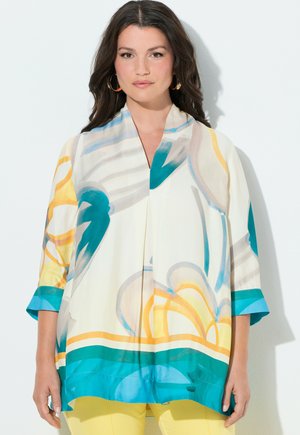 Mujer con cabello largo y oscuro, que lleva pendientes de aro dorados y una blusa suelta de manga larga con patrones florales abstractos en amarillo, azul y verde azulado.