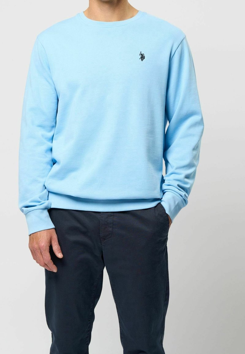 Vaaleansininen collegepaita pyöreällä kaula-aukolla ja pitkillä hihoilla, jossa pieni musta logo rinnassa. Pehmeä puuvillainen pinta.