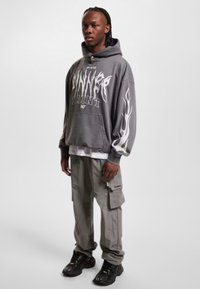 Grå oversized hettegenser med hvite flammegrafikker, brukt over en hvit skjorte. Kombinert med lysegrå cargo-bukser og svarte sneakers.