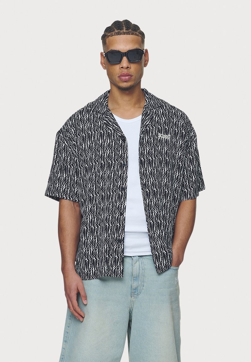 Camisa de manga curta com padrão preto e branco, com design de botões, combinada com um top branco e calças de ganga largas em azul claro. Óculos de sol pretos.