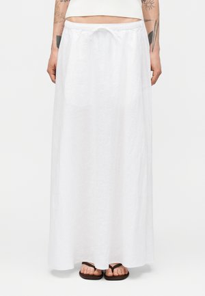 Kvinde iført et langt hvidt linnednederdel med snøre i taljen og sorte flip-flops, stående mod en ensfarvet baggrund.