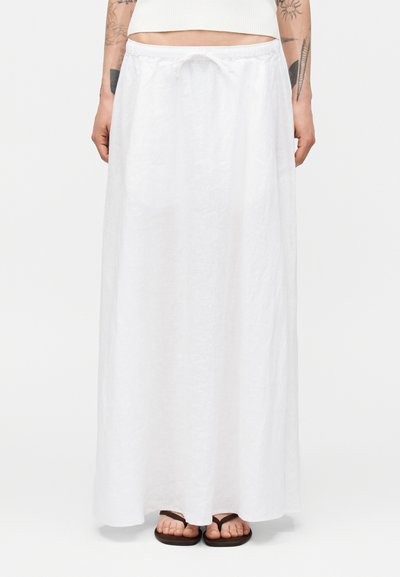 Femme portant une longue jupe en lin blanc avec une taille à cordon et des tongs noires, debout devant un fond uni.