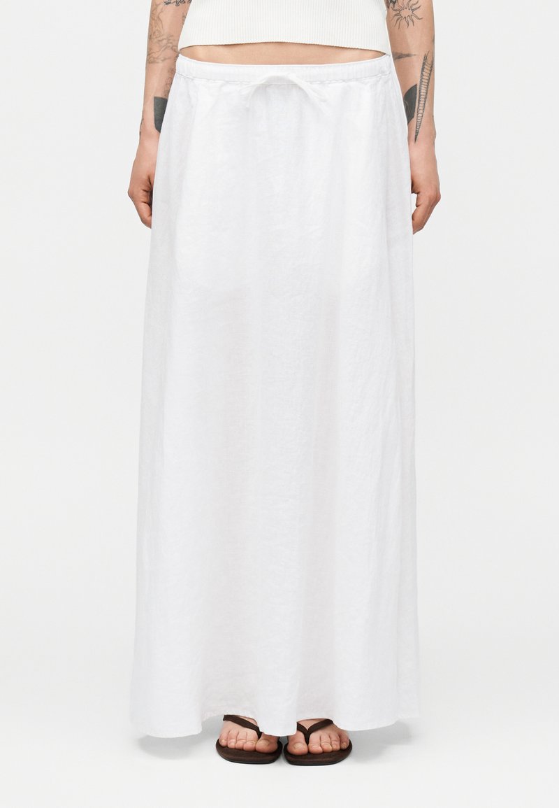 Γυναίκα που φοράει μακριά λευκή λινή φούστα με κορδόνι στη μέση και μαύρα σαγιονάρες, στέκεται μπροστά σε απλό φόντο.