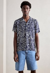 Camisa azul-marinho de manga curta com padrões abstratos brancos, com botões à frente, combinada com calções de ganga claros. Textura suave, ajuste descontraído.
