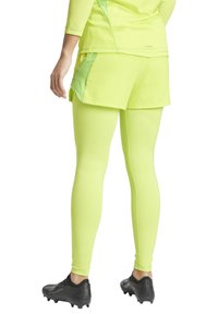 Neon gele sportoutfit, bestaande uit strakke leggings en een losse top, met gaasdetails en contrasterende naden, gecombineerd met zwarte sportschoenen.
