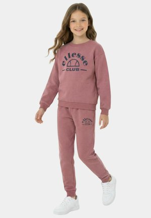 Jeune fille souriante, portant un sweat-shirt Club ellesse rose poussiéreux et un jogging assorti avec des baskets blanches, posant devant un fond uni.