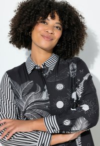 Sort skjorte med knapper og stribede ærmer, prydet med kontrasterende hvide blomstermønstre og solmotiver. Blødt stof med klassisk kravedesign.