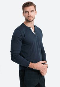 Mörkblå långärmad henley-tröja med en vit krage och träknappar. Tyget är slätt med en figurnära passform, i kombination med rutiga byxor.