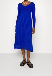 Robe en tricot bleue avec des manches longues, un corsage texturé et une jupe côtelée présentant un ourlet légèrement évasé. Associée à des sandales noires.