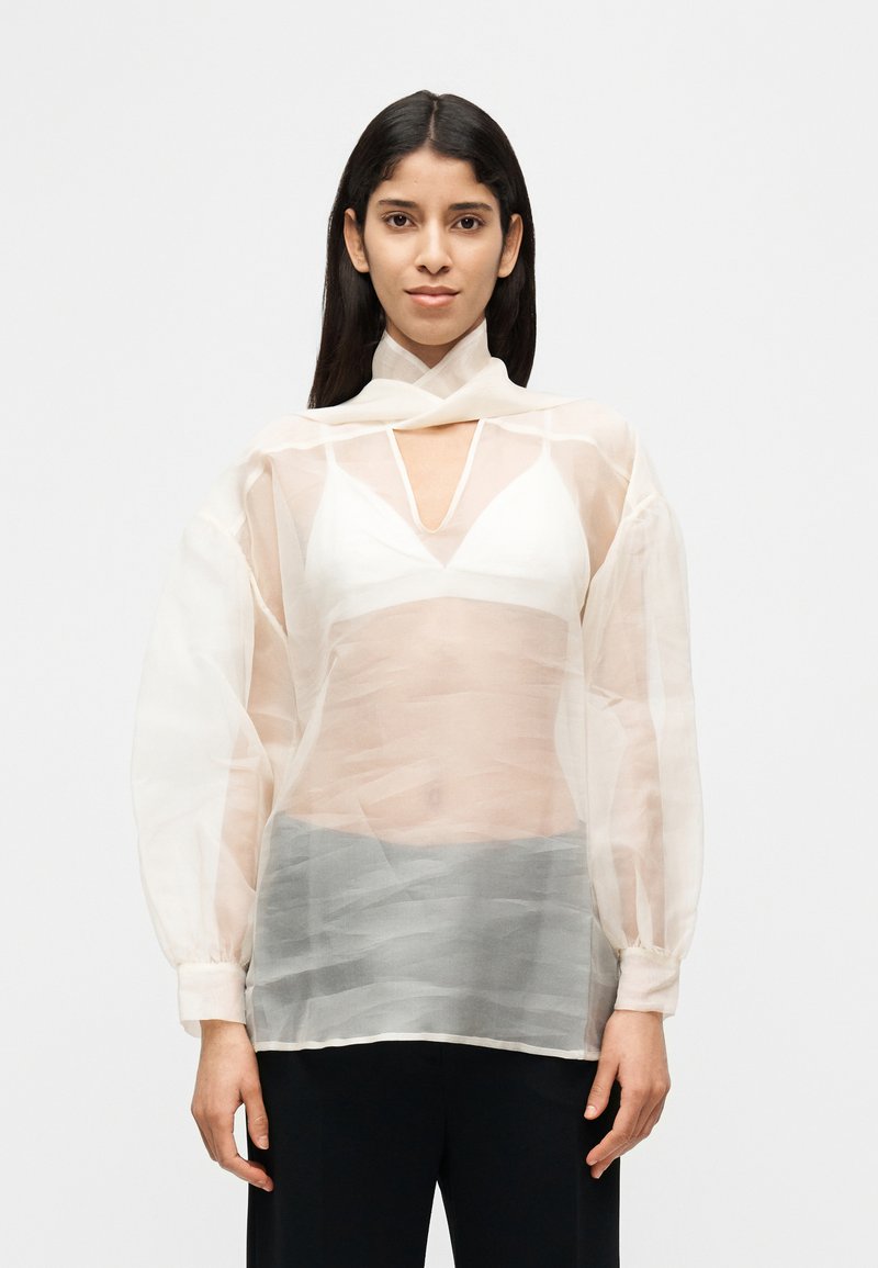 Ελαφρύ, διάφανο πουκάμισο σε απαλό, ανοιχτό χρώμα με V-λαιμόκοψη και μακριά, μπαλόν μανίκια. Διαθέτει στρωματοποιημένο, υφασμάτινο υλικό με υφή για βάθος.