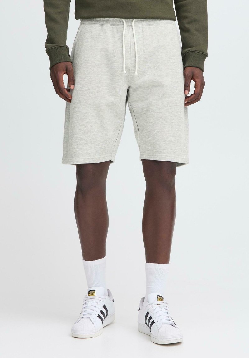 Pantalones cortos de algodón grises con cintura elástica y cordón ajustable. Modelo llevando calcetines blancos y zapatillas de deporte con rayas negras. Diseño simple y casual.
