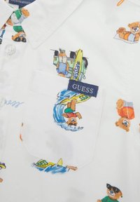 Camicia bianca a maniche corte con orsetti cartoon colorati in varie attività, con un'etichetta blu "GUESS" sulla tasca del petto.