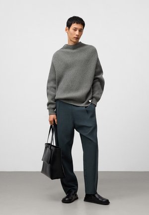 Jasnoszary sweter z prążkowanej dzianiny, luźny krój, w parze z ciemnozielonymi spodniami o szerokich nogawkach. Modelka trzyma czarną skórzaną torbę, nosząc czarne buty.