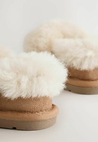 Tan suede tossut, joissa on pörröinen valkoinen turkisreunus, pyöreä muoto, teksturoitu pohja ja pehmeä sisus mukavuutta varten.