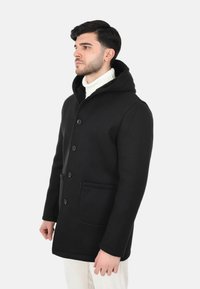 Cappotto nero in lana con cappuccio, caratterizzato da una chiusura frontale con bottoni, due tasche laterali e una texture liscia. Indossato sopra un dolcevita bianco.