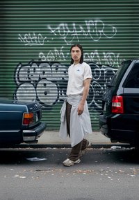 Wit T-shirt met blauw logo, grijs-wit gestreepte wikkel, bruine broek en lichte beige sneakers. Geplaatst tegen een groene graffiti-muur.