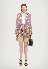 Blazer à motif floral violet avec des accents verts, chemise blanche, shorts beiges et sac à main noir. Des sandales noires à brides complètent le look.