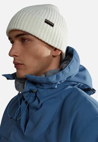 Giovane uomo che indossa un berretto di lana bianco e un giubbotto invernale blu con colletto alto e cappuccio regolabile, mostrato di profilo.