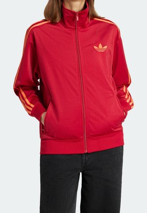 Person trägt eine rote Trainingsjacke mit durchgehendem Reißverschluss, orangenen Streifen an den Ärmeln und einem Logo auf der Brust, kombiniert mit schwarzer Hose, Hände in den Taschen.