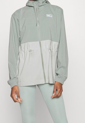 Kobieta w jasnozielonej, zapinanej na pełny zamek bluzie z kapturem i ściągaczem w talii oraz dopasowanych legginsach na jednolitym tle.