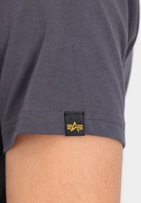 Siva majica s kratkimi rokavi, ki se ponaša z gladko teksturo in črnim oznako z zlatim logotipom na robu rokava.