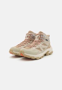 Beige vandringsskor med meshtyg och mockadetaljer, klara orange snören, texturerade gummisulor och mid-top design för stöd av ankeln.