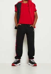 Červené tričko s černým logem, spárované s černými tepláky s červenými akcenty. Černé boty s červenými detaily. Mikina přehozená přes ramena.