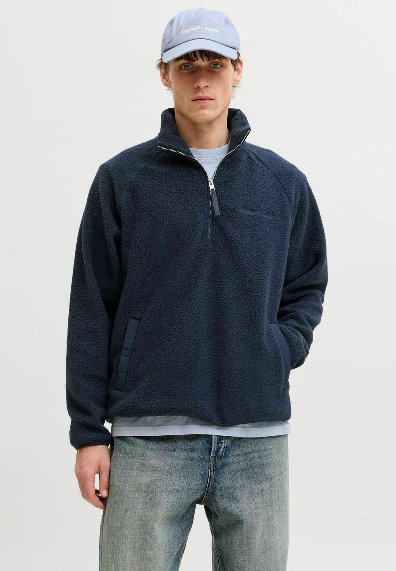 Bărbat tânăr purtând o șapcă albastru deschis, pulover fleece bleumarin cu fermoar pe jumătate, cămașă gri deschis și blugi albaștri decolorați, stând în picioare pe un fundal simplu.