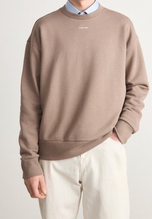 Person trägt einen braunen Calvin Klein Sweatshirt über einem hellblauen Hemd mit Kragen und offwhite-farbenen Hosen, mit einer Hand in der Tasche und der anderen seitlich.