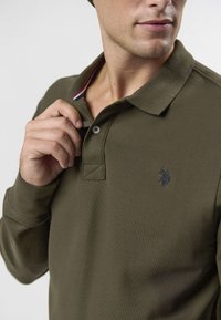 Olivgrön långärmad pikétröja med treknappad knäppning, stickad textur och broderad logga på bröstet. Krage med ränder som accent.