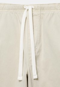 Pantalon beige clair avec ceinture élastique et long cordon blanc noué en un simple nœud au centre devant.