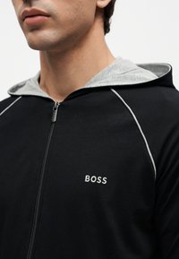 Uomo che indossa una felpa nera con zip, fodera grigia nel cappuccio e la scritta "BOSS" ricamata sul petto.