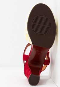 Chaussure à talon haut en daim rouge avec bride à la cheville, montrant une semelle texturée avec le nom de la marque «Chie Mihara» et un motif à pois jaunes sur le côté de la chaussure.