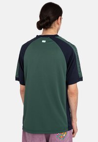 Camisa deportiva verde y azul marino con mangas raglán, que cuenta con una etiqueta en la parte de atrás y una textura suave. Destaca por su diseño de bloques de color.
