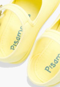Zapatillas de lona amarillas con una textura suave, con un diseño redondeado, una correa en la parte superior y "Pisamonas" impreso en el interior.