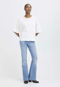 Kvinde står med hænderne bag ryggen iført en løs hvid t-shirt, lyseblå flared jeans og sorte hæle på en hvid baggrund.