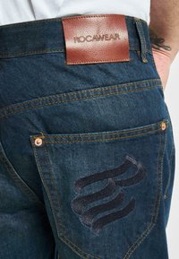Jeans de mezclilla oscuro con una etiqueta de cuero ROCWEAR. Presentan herrajes dorados y un diseño de espiral negra bordado en el bolsillo trasero.