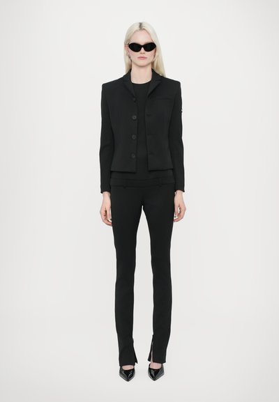 Black tailored blazer with button front, paired with fitted black pants featuring split hems. Model wears black sunglasses and heels.
