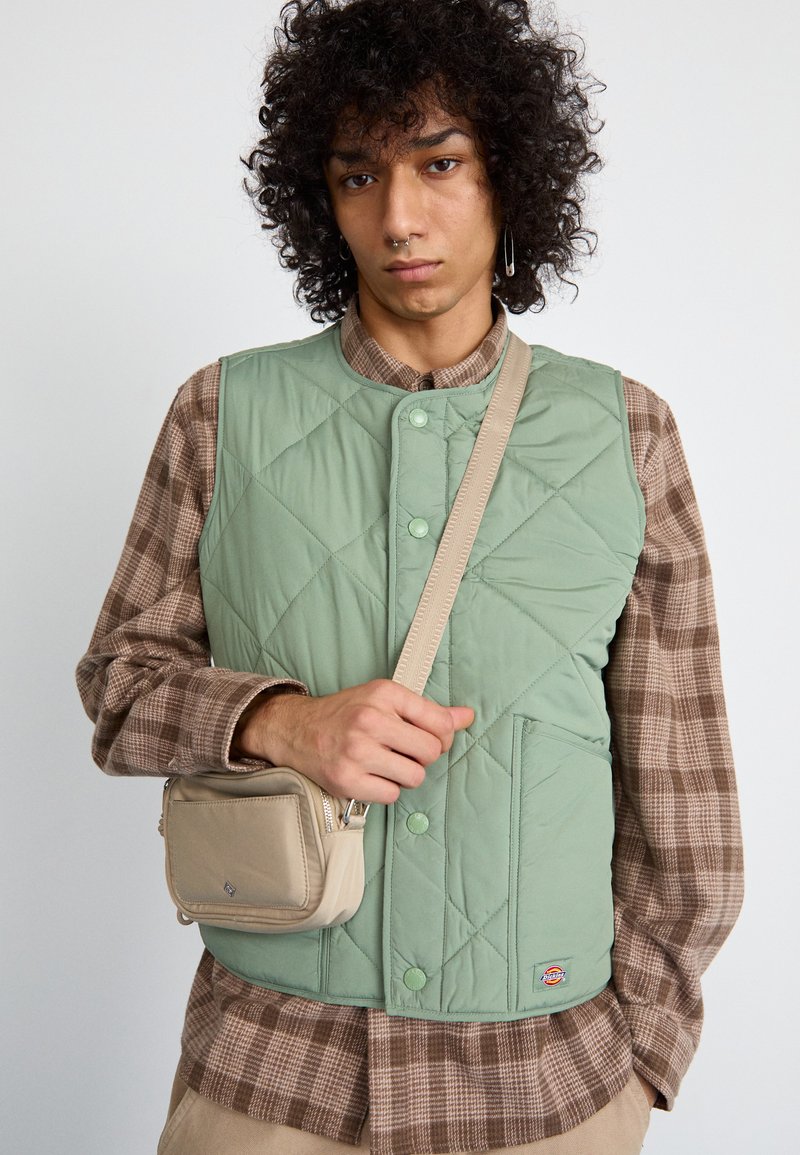 Lysegrøn, ærmeløs quilted vest med rund halsudskæring, trykknaplukninger og frontlommer; parret med en brunternet skjorte og en lille beige taske.