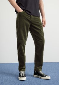 Groene ribfluwelen broek met een geribbeld textuur, voorzien van zijzakken en een rechte snit. Gecombineerd met zwarte hoge sneakers en witte veters.