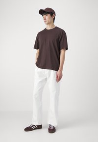 Donkerbruine katoenen T-shirt, witte relaxed-fit broek en bruine sneakers met witte strepen. Gecombineerd met een bijpassende bruine pet.